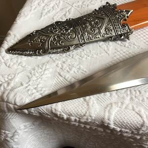 Puede incluir: Una espada decorativa de plata y oro con un mango detallado y una hoja afilada. La espada est&aacute; sobre una superficie de tela blanca.
