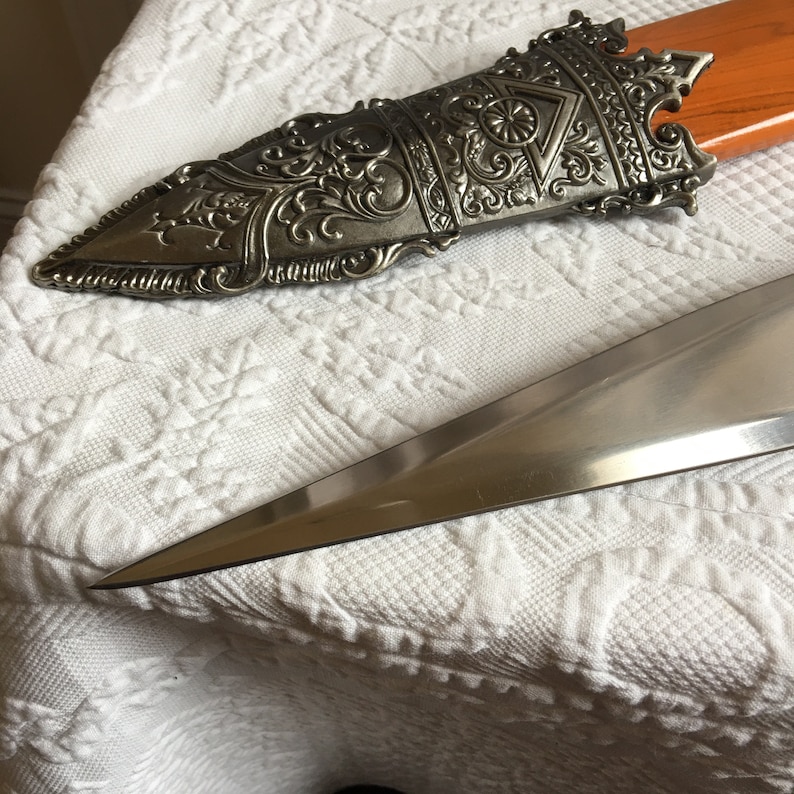 Puede incluir: Una espada decorativa de plata y oro con un mango detallado y una hoja afilada. La espada est&aacute; sobre una superficie de tela blanca.