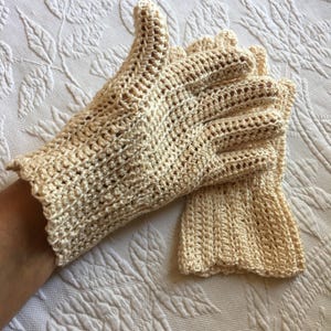 May include: A pair of cream-colored, crocheted gloves. The gloves have a delicate, openwork design and a scalloped edge at the wrist. The gloves are made of a soft, textured yarn and appear to be handmade.