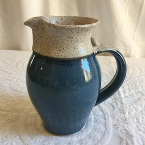 Puede incluir: Una jarra de cerámica azul y blanca con un esmalte moteado. La jarra tiene un cuerpo redondeado y un asa.