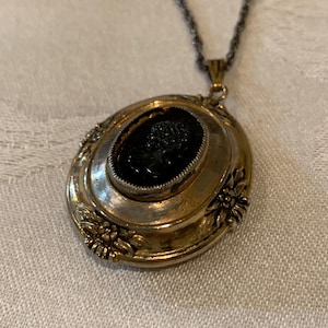 May include: A vintage gold-toned oval pendant necklace with a black cameo. The pendant has a detailed floral design around the edges and a dark chain. The cameo features a detailed profile.