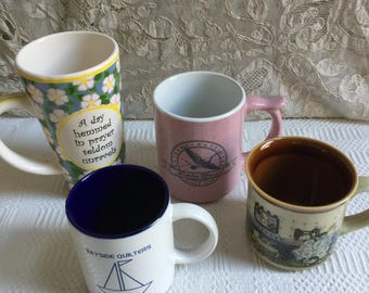 May include: Four ceramic mugs with various designs. One mug has a floral pattern and the text "A day hemmed in prayer seldom unravels." Another is pink with a logo. A third is white with a blue interior and a sailboat graphic. The last mug has a brown interior.