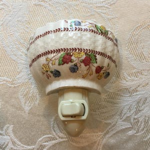 May include: A ceramic night light with a floral design. The night light has a cream-coloured base with a switch and a scalloped bowl-shaped top. The top is decorated with a band of red, yellow, and blue flowers and berries.