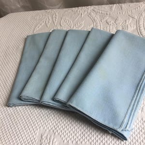 May include: A set of five folded light blue cloth napkins. Each napkin is rectangular in shape with a smooth, woven texture and finished edges. The napkins are stacked in a fan-like arrangement, displaying a consistent solid pastel blue color.