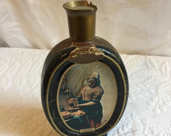 May include: A vintage black and gold decanter with a worn label featuring a painting of a woman pouring milk. The label reads "A Maid Serving Milk".
