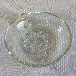 Pode incluir: Tigela de vidro transparente com uma pega e um desenho floral no centro. A tigela é redonda e transparente. A pega está presa à lateral da tigela. Adequada para servir snacks ou sobremesas.