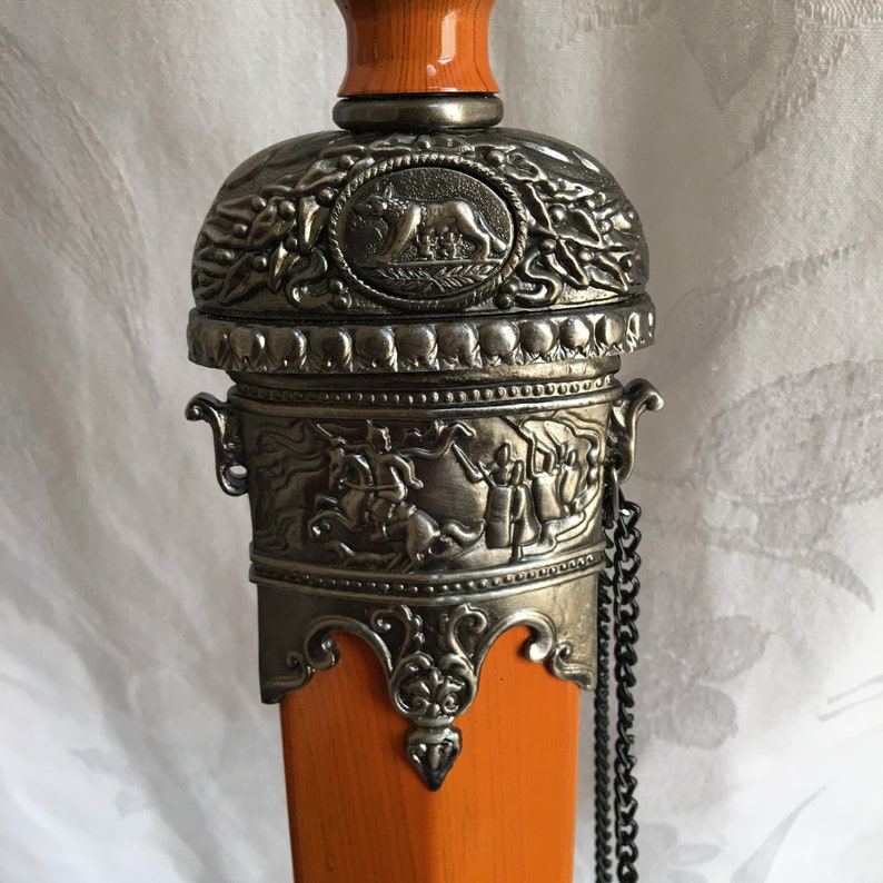 Puede incluir: Primer plano de una vaina de espada decorativa con mango de madera naranja y parte superior de metal plateado. La parte superior presenta un dise&ntilde;o de le&oacute;n y detalles intrincados.