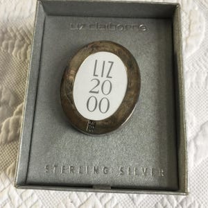May include: A Liz Claiborne sterling silver oval-shaped pendant with the text "LIZ 2000" on a white background. The pendant is in a gray box with "STERLING SILVER" printed on it.