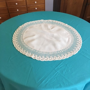 Puede incluir: Un tapete redondo de color crema con un borde de ganchillo, colocado sobre un mantel turquesa. El centro del tapete es de tela lisa y uniforme. El borde de ganchillo presenta un delicado patrón repetitivo. La mesa está en una habitación con muebles de madera.