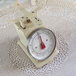 Puede incluir: Balanza de cocina vintage de color crema con esfera circular y aguja roja. La balanza tiene una capacidad de 500 gramos. La balanza presenta un cuerpo color crema y una esfera metálica con números y marcas negras. La balanza tiene un diseño retro.