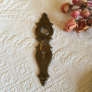 May include: A brass keyhole cover with an ornate design. The cover has a keyhole in the center and a round opening at the top.