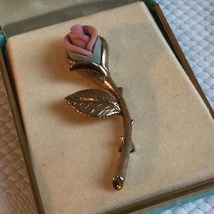 May include: A vintage rose brooch. The brooch features a pink and light blue rose bud with a gold-coloured stem and leaf. The brooch is displayed in a vintage jewellery box with a light beige interior.