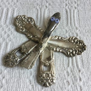 May include: A whimsical sculpture made from antique silverware. The arrangement features spoons and forks, with googly eyes added to a knife, creating a unique, artistic piece. The silverware has ornate detailing and a vintage aesthetic.