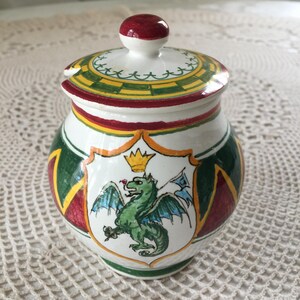 May include: A white ceramic jar with a lid, featuring a hand-painted design. The jar has a green dragon emblem on the front, with green, yellow, and red accents. The lid is topped with a red knob.