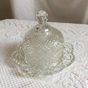 May include: A clear glass butter dish with a domed lid and a scalloped base. The dish has a cut glass design.