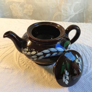 Op de afbeelding: Een bruine keramische theepot met bijpassende deksel. De theepot is versierd met witte, blauwe en groene bloemaccenten. Het deksel ligt opzij en onthult de binnenkant van de theepot.