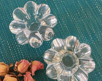 May include: Two clear glass candle holders shaped like flowers with six petals each. The candle holders are on a green textured surface.