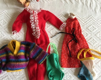 May include: A selection of vintage doll clothes, including a red jumpsuit with white lace trim, a red floral dress, a green swimsuit, a striped sweater, and other accessories. The items are arranged on a white textured surface.