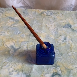 Peut inclure: Un encrier en verre bleu avec une plume à tremper en bois à l'intérieur. L'encrier a un bouchon en liège et repose sur une surface à motifs bleus et blancs.