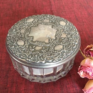 May include: A silver-plated glass trinket box with a floral design on the lid. The box is round and has a clear glass base.