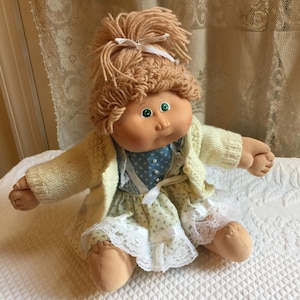 Könnte beinhalten: Eine Vintage Cabbage Patch Kid Puppe mit blonden Garnhaaren, grünen Augen und einer rosa Schleife. Die Puppe trägt einen weißen Strickcardigan, ein blau-weißes Blumenkleid und einen weißen Spitzenbesatz.