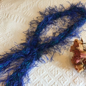 May include: A blue and teal fuzzy scarf with long fringe. The scarf is laid out on a white textured surface.