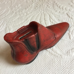 May include: Red ceramic boot shaped piggy bank with a slot on the top for coins. The boot has a textured surface and a black heel.