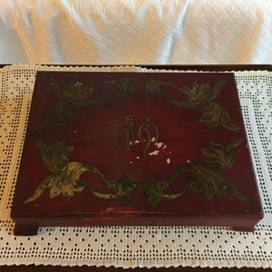 Caja antigua. Pintura roja craquelada con diseños de hojas en espiral en oro envejecido. Monograma MOR en el centro. Cuatro patas en las esquinas. Caja encantadora.