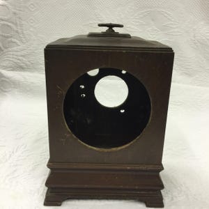 May include: A dark brown wooden clock case with a square shape and a circular opening for the clock face. It has a small handle on top and sits on a rectangular base. The clock case is likely vintage or antique.