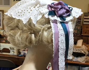 Puede incluir: Bonnet de encaje blanco con una cinta morada y turquesa y un acento de flor morada.