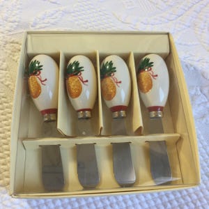 May include: A set of four metal spreaders with white ceramic handles, each featuring a painted yellow pineapple graphic with green leaves and a red ribbon detail. The spreaders are arranged side by side in a light beige cardboard display box.