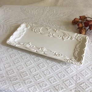 以下が含まれることがあります： 縁に装飾的な浮き彫りのスクロール模様が施された、長方形のオフホワイトのセラミックトレイ。 トレイはわずかにテクスチャ加工された表面とシンプルなエレガントなデザインです。 背景にはドライフラワーがあります。