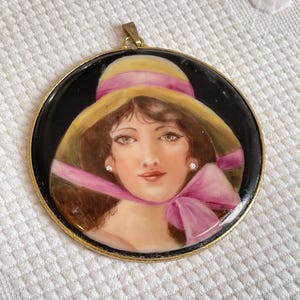 May include: A round porcelain pendant with a gold rim, featuring a painted portrait of a woman wearing a yellow and pink hat with a pink bow. The woman has brown hair, brown eyes, and is wearing pearl earrings.