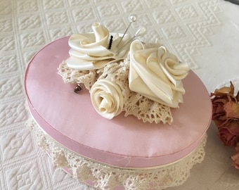 May include: A pink round box with a white lace trim and a white satin flower arrangement on top. The flower arrangement includes three white satin roses and a few pearl-headed pins.