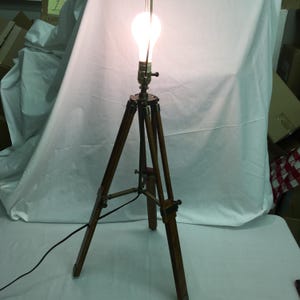 May include: A vintage-style wooden tripod floor lamp with a large, illuminated light bulb. The lamp features a dark brown wooden tripod base with brass-colored metal accents. The bulb emits a bright, warm light, and a black power cord is visible.
