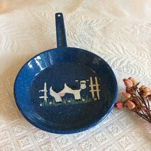 May include: Blue enamel frying pan with a white cow illustration and a pink udder. The cow is standing near a white fence with green grass and flowers.
