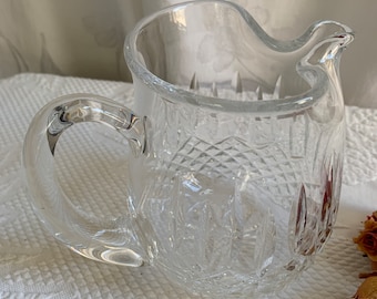 May include: A clear glass pitcher with a cut glass design. The pitcher has a handle and a spout.