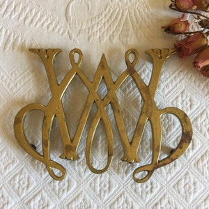 Puede incluir: Un salvamanteles de latón vintage con un diseño ornamentado con las letras entrelazadas "WM". El salvamanteles tiene un tono dorado con signos de edad y desgaste, y se coloca sobre una superficie blanca texturizada. Flores secas en el fondo.