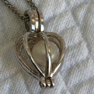 Puede incluir: Collar con colgante en forma de corazón plateado. El colgante presenta un diseño de jaula que contiene una sola perla redonda blanquecina. La cadena del collar también es plateada y visible en la parte superior. El fondo es una tela blanca texturizada.