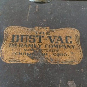 Puede incluir: Primer plano de un objeto metálico marrón vintage con una etiqueta dorada. La etiqueta dice "THE DUST-VAC" encima de "THE RAMEY COMPANY MANUFACTURERS CHILLICOTHE, OHIO, U.S.A."