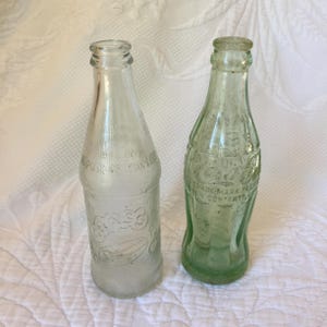 Puede incluir: Dos botellas de refresco de vidrio vintage. Una es transparente y la otra es verde. Ambas tienen letras en relieve en el lateral. La botella verde tiene el texto "Coca-Cola" y "Trademark Registered".