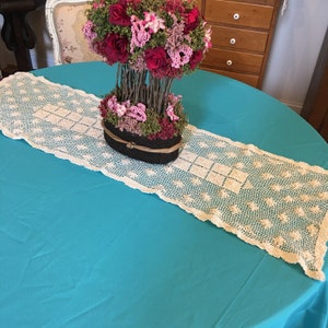 Op de afbeelding: Een turquoise tafelkleed met een witte gehaakte kantloper. Een bloemstuk met roze en rode bloemen staat in een bruine geweven mand op de loper.