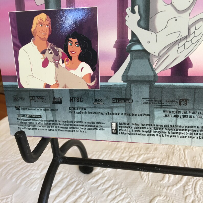 May include: A VHS tape case with a pink, purple, and blue background. The front of the case features a cartoon image of a man, woman, and a goat. The text on the case reads "NTSC Extended Play" and "This Laserdisc is Extended Play".