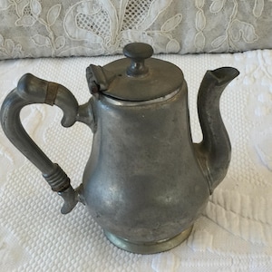 Puede incluir: Una pequeña tetera antigua de peltre con asa y boquilla curvadas. La tapa tiene un pequeño pomo y el asa está sujeta con una banda metálica decorativa. La tetera está sobre una superficie blanca texturizada.