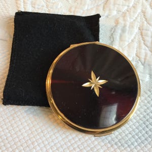 May include: A vintage compact with a dark, glossy surface and a gold starburst design. The compact has a gold-colored rim and is accompanied by a black velvet pouch. The compact is a classic beauty accessory.