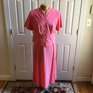 Może przedstawiać: Różowa sukienka dwuczęściowa z krótkim rękawem i spódnicą do kolan. Góra ma kołnierzyk typu mandarynka i zapięcie na guziki z przodu.