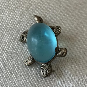 May include: A silver-toned turtle brooch with a large, oval, light blue cabochon center. The turtle's shell is the blue stone, and the head, feet, and tail are detailed metal. The brooch is a decorative accessory.