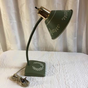 May include: A desk lamp with a sage green base and a matching conical shade featuring white leaf patterns. The lamp has a flexible dark green neck and a polished gold-tone metal connector at the top of the shade. A beige power cord is attached.