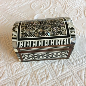 Puede incluir: Una pequeña caja de madera con un patrón geométrico blanco y negro incrustado con nácar. La caja tiene una tapa curva y está cerrada.