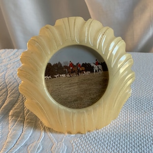 Puede incluir: Un marco de fotos festoneado de color crema con una abertura circular. El marco contiene una fotografía de personas a caballo, acompañadas de perros, en un campo. El marco tiene un diseño texturizado y ondulado en el borde.